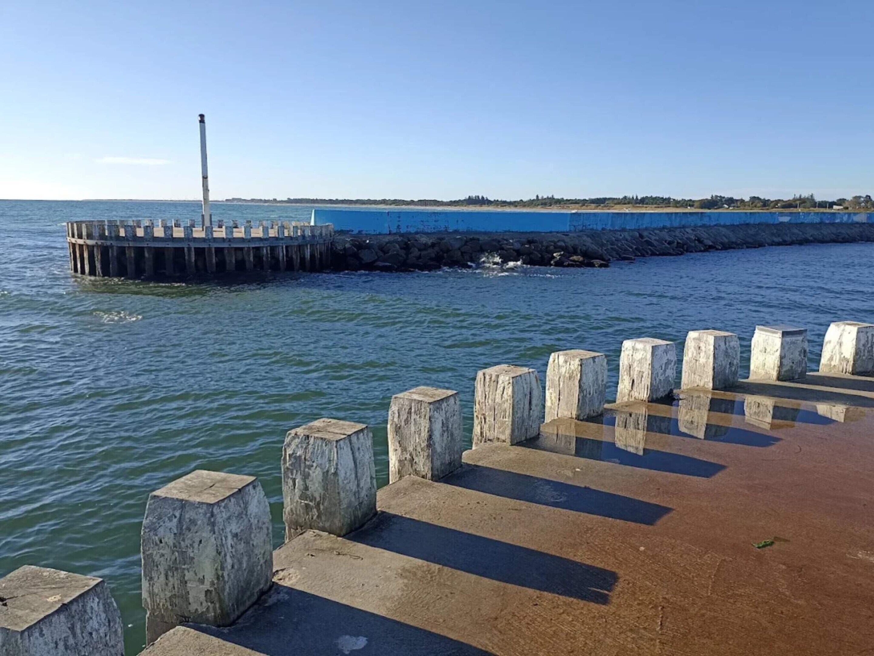Eine Steg mit Betonstützen und einem Leuchtturm im Hintergrund.