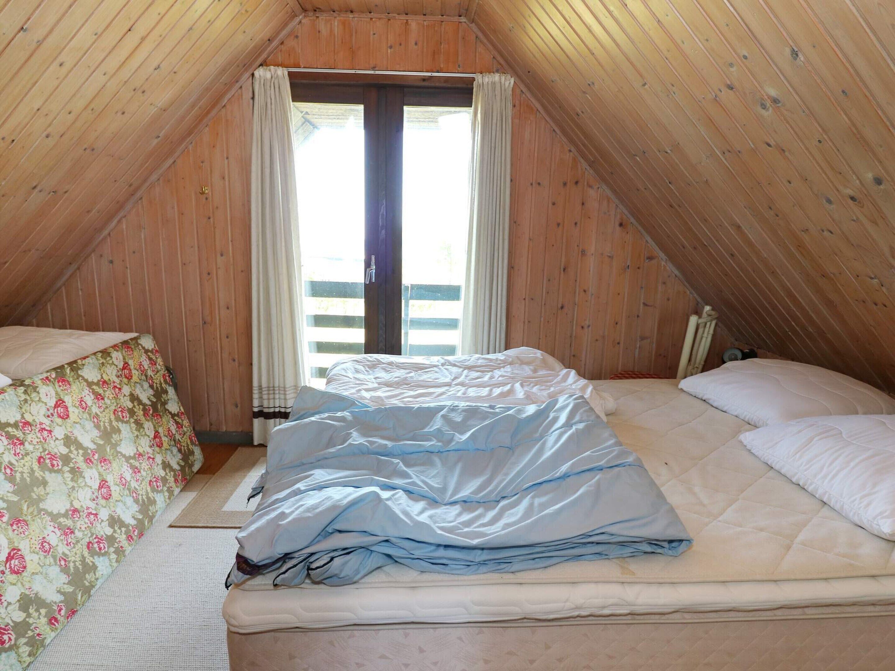 Een kamer met een schuine kap, een bed met matras en kussens, een raam met gordijnen en een houten vloer.