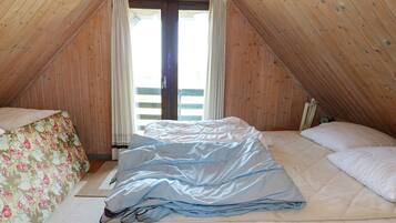 A room with a sloped ceiling, a bed with a mattress and pillows, a window with curtains, and a wooden floor.