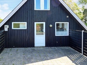 A dark wooden building with a white door, two windows, and a small outdoor area with a paved floor. - 6 Person Holiday Home in Hadsund (Hadsund)
