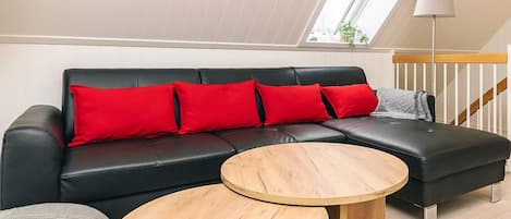 A modern loft-style room with a black leather sofa, red cushions, and two wooden side tables.