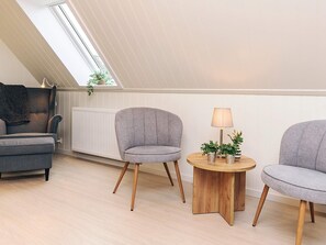 Un dormitorio tipo loft moderno con techo inclinado, un sofá gris, dos sillones, una mesita de madera con plantas y un radiador.