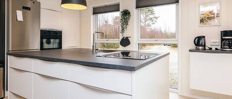 A modern kitchen with a central island, a built-in oven, a refrigerator, and a hanging plant.
