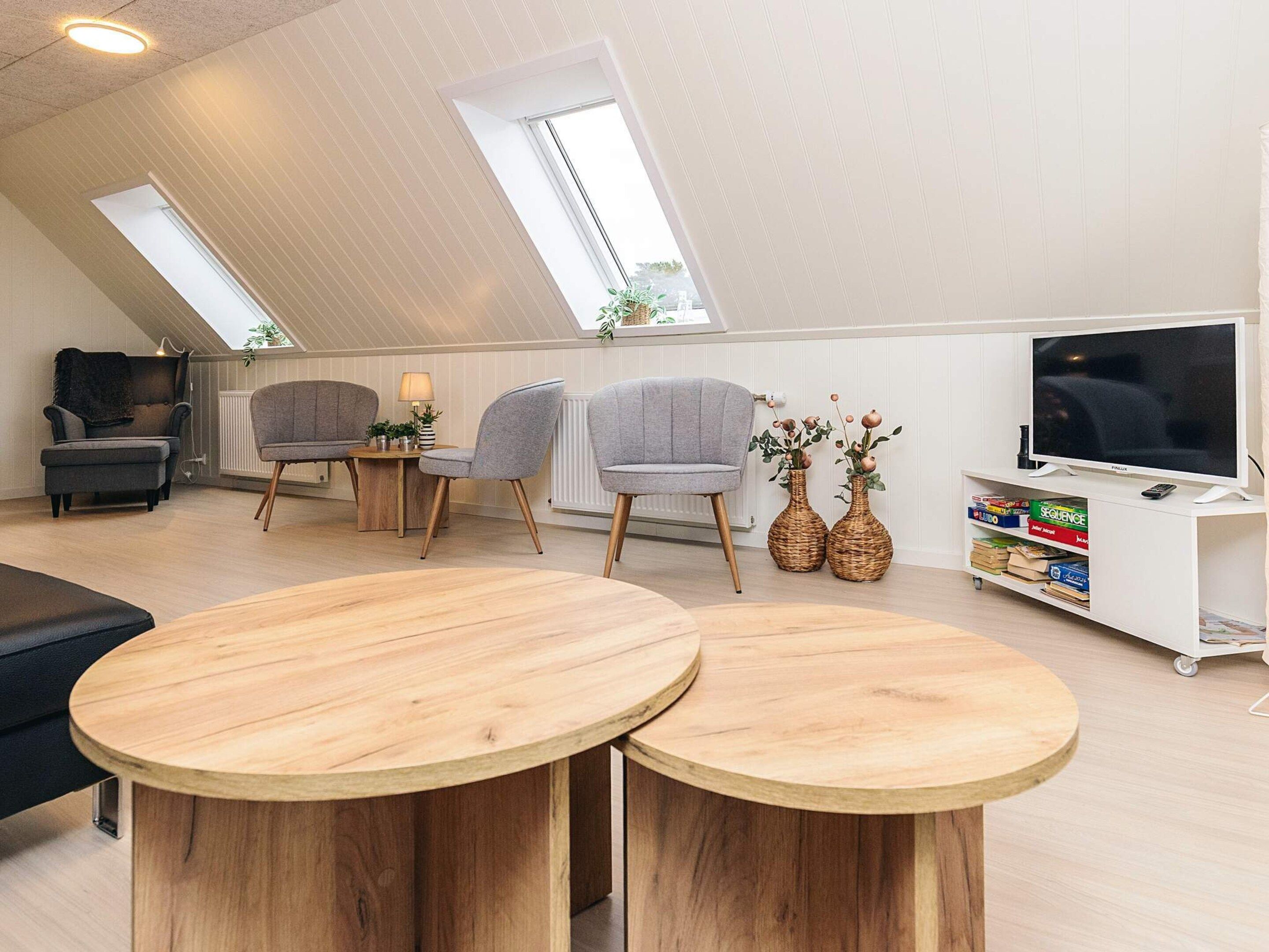 A loft-style room with a sloped ceiling, two round wooden tables, grey upholstered chairs, a television on a white stand, and a bookshelf.