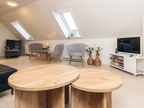 Una habitación estilo loft con techo inclinado, dos mesas redondas de madera, sillas tapizadas de color gris, un televisor sobre un soporte blanco y una estantería.