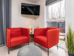 Una sala moderna con dos sillones rojos, un televisor de pantalla plana, una mesita auxiliar con una lámpara y una planta en maceta.