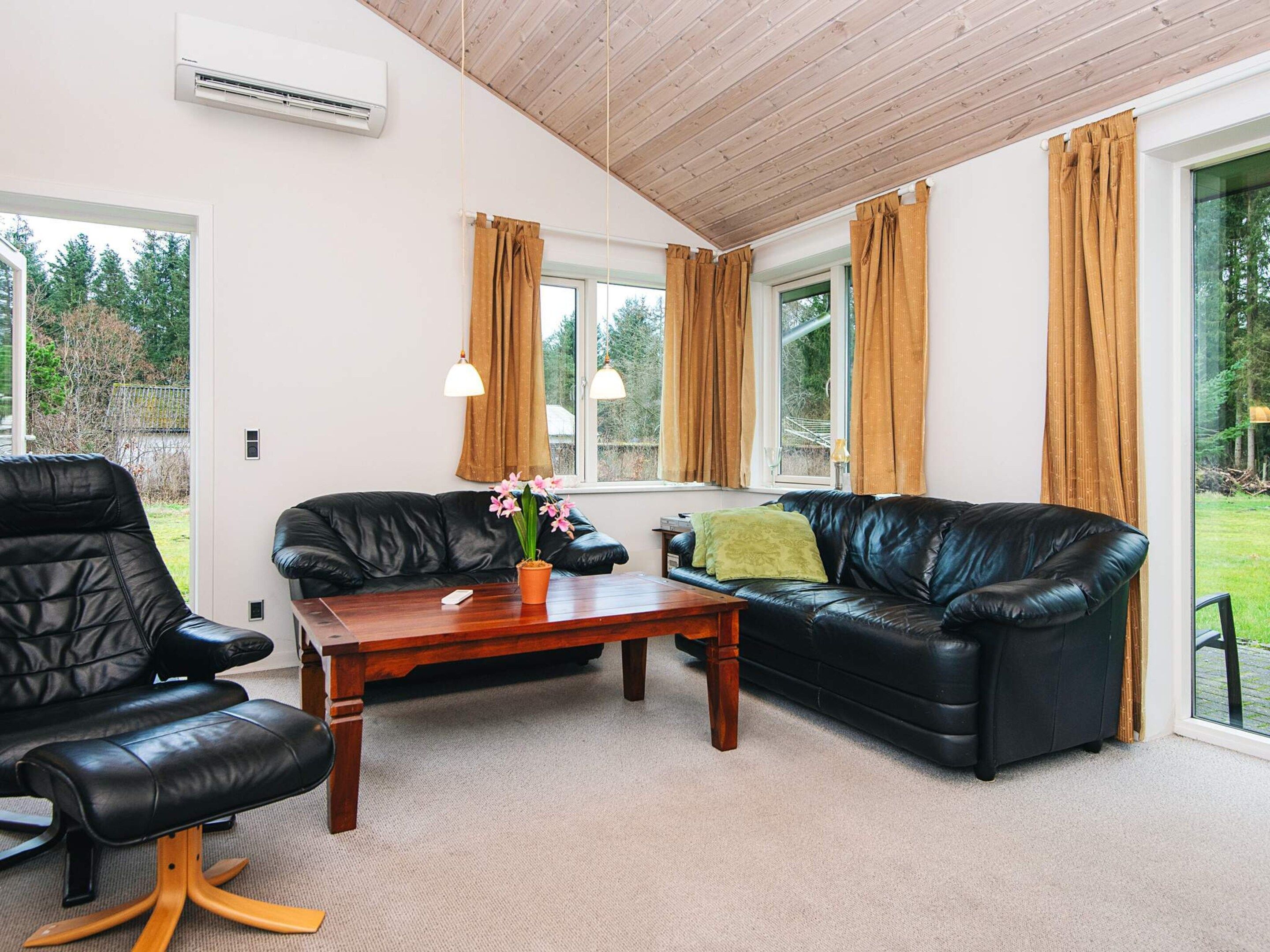 A modern living room with a black leather sofa, a wooden coffee table, and a black leather armchair. There is a window with curtains, a ceiling fan, and an air conditioning unit.