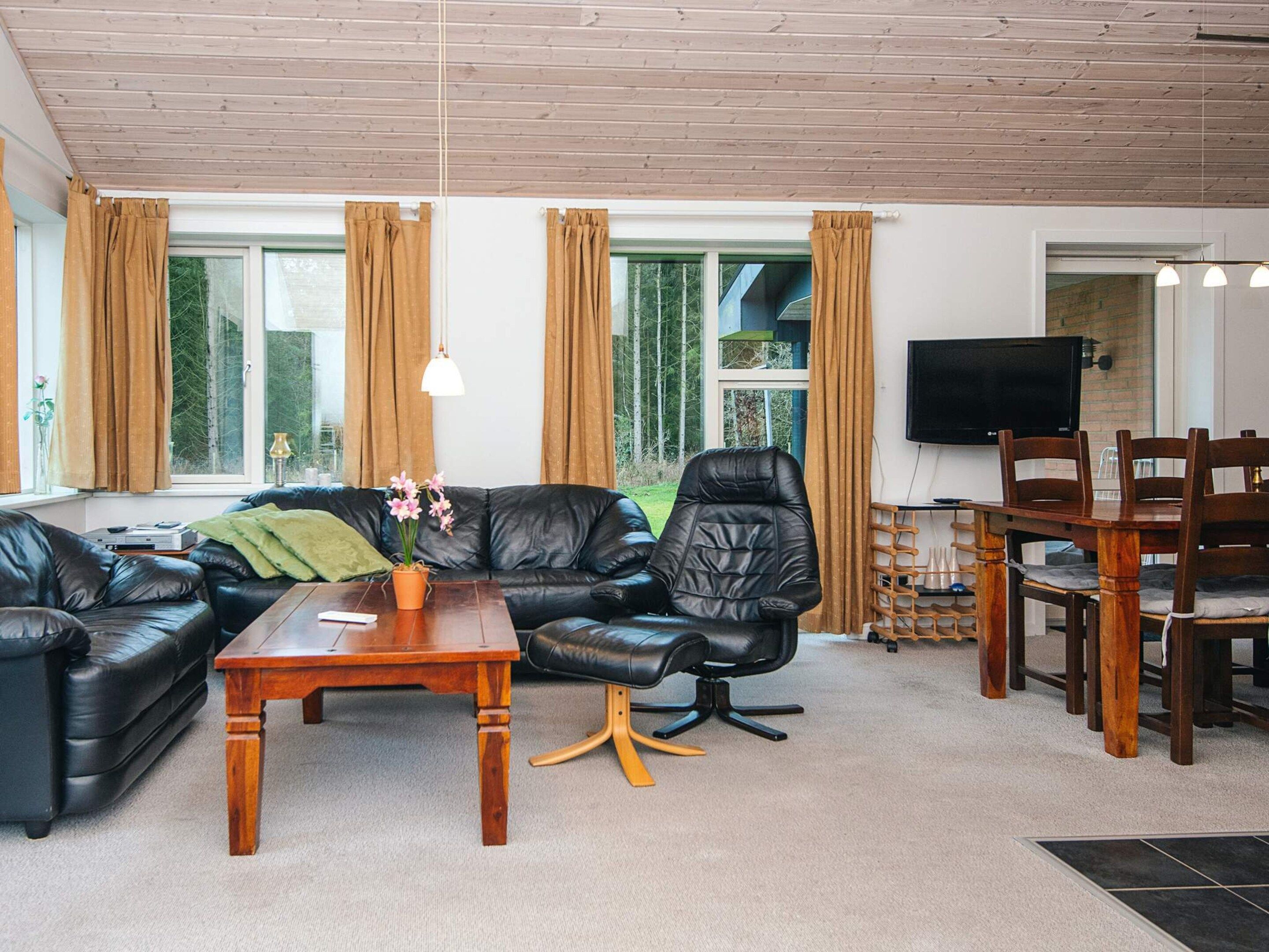 Una sala de estar moderna con un sofá de cuero negro, una mesa de centro de madera y un sillón de cuero negro. Se observa una zona de comedor con una mesa y sillas de madera. Amplias ventanas con cortinas son visibles, y un televisor de pantalla plana está montado en la pared.