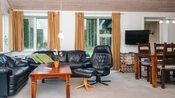 Una sala de estar moderna con un sofá de cuero negro, una mesa de centro de madera y un sillón de cuero negro. Se observa una zona de comedor con una mesa y sillas de madera. Amplias ventanas con cortinas son visibles, y un televisor de pantalla plana está montado en la pared.