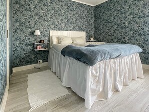 A bedroom with a bed, white bedding, a blue blanket, a nightstand, a lamp, and a floral wallpaper.