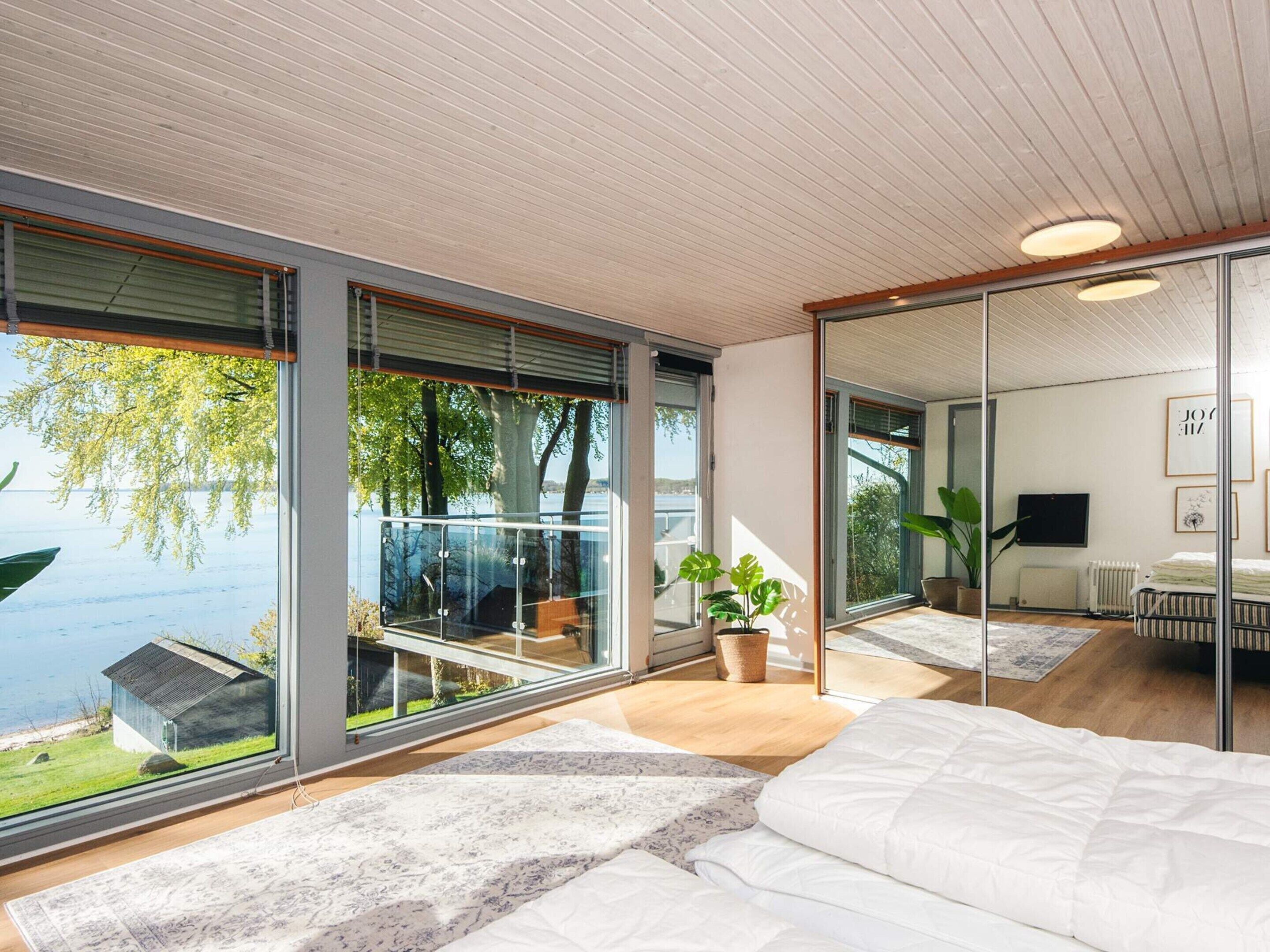 A modern bedroom with large windows, a bed, a plant, and a view of a lake.