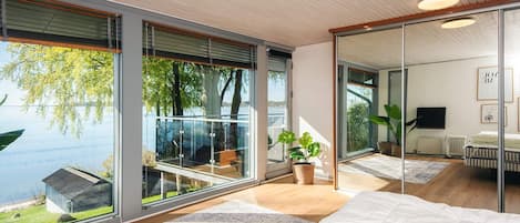 A modern bedroom with large windows, a bed, a plant, and a view of a lake.