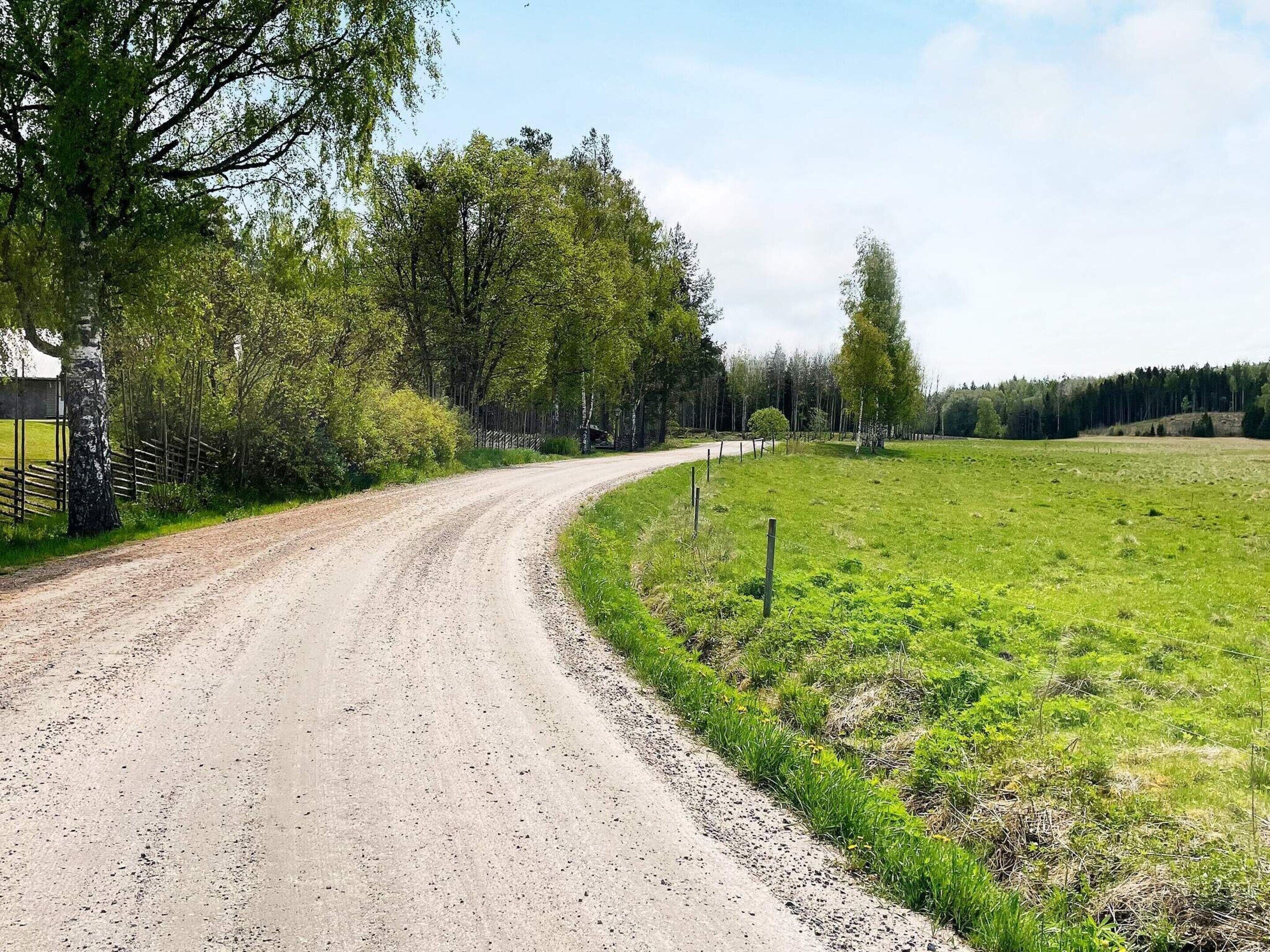 A rural road curving through a green field with trees on one side and a fence on the other.