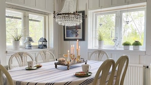 A dining area with a striped tablecloth, six chairs, a chandelier, candles, and potted plants on the windowsill. - 7 Person Holiday Home in Klassbol (Klässbol)