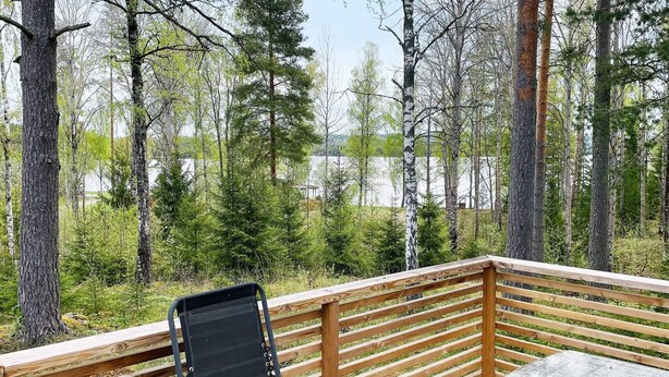 A wooden deck with a black chair and a glass table, surrounded by trees and a view of a body of water. - 7 Person Holiday Home in Klassbol (Klässbol)