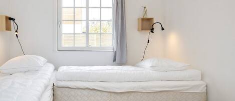 A room with two single beds, a window with curtains, a wooden floor, and two wall-mounted lamps.