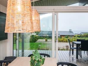 A modern outdoor dining area with a wooden table and chairs, a hanging wicker light fixture, and a view of a garden and buildings across a body of water.