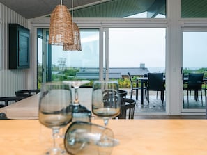 A dining area with a wooden table and chairs, a wicker pendant light, and a view of a building and beach through large glass doors. - 6 Person Holiday Home in Ronde (Ronde)