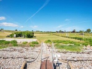 Paisatge rural amb un camí de grava que condueix a un pont de fusta sobre un rierol, envoltat de verdura i un cel serè.