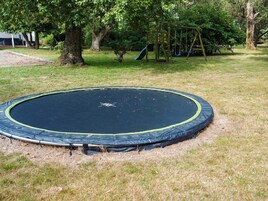 Un trampolín en un área con césped, árboles y un parque infantil al fondo.