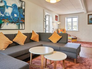 Un salon moderne avec un canapé d’angle, deux tables rondes, un fauteuil coloré et un grand tableau abstrait.