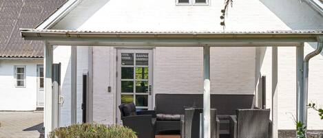 A covered outdoor seating area with wicker furniture, a wooden deck, and a white building with a window.