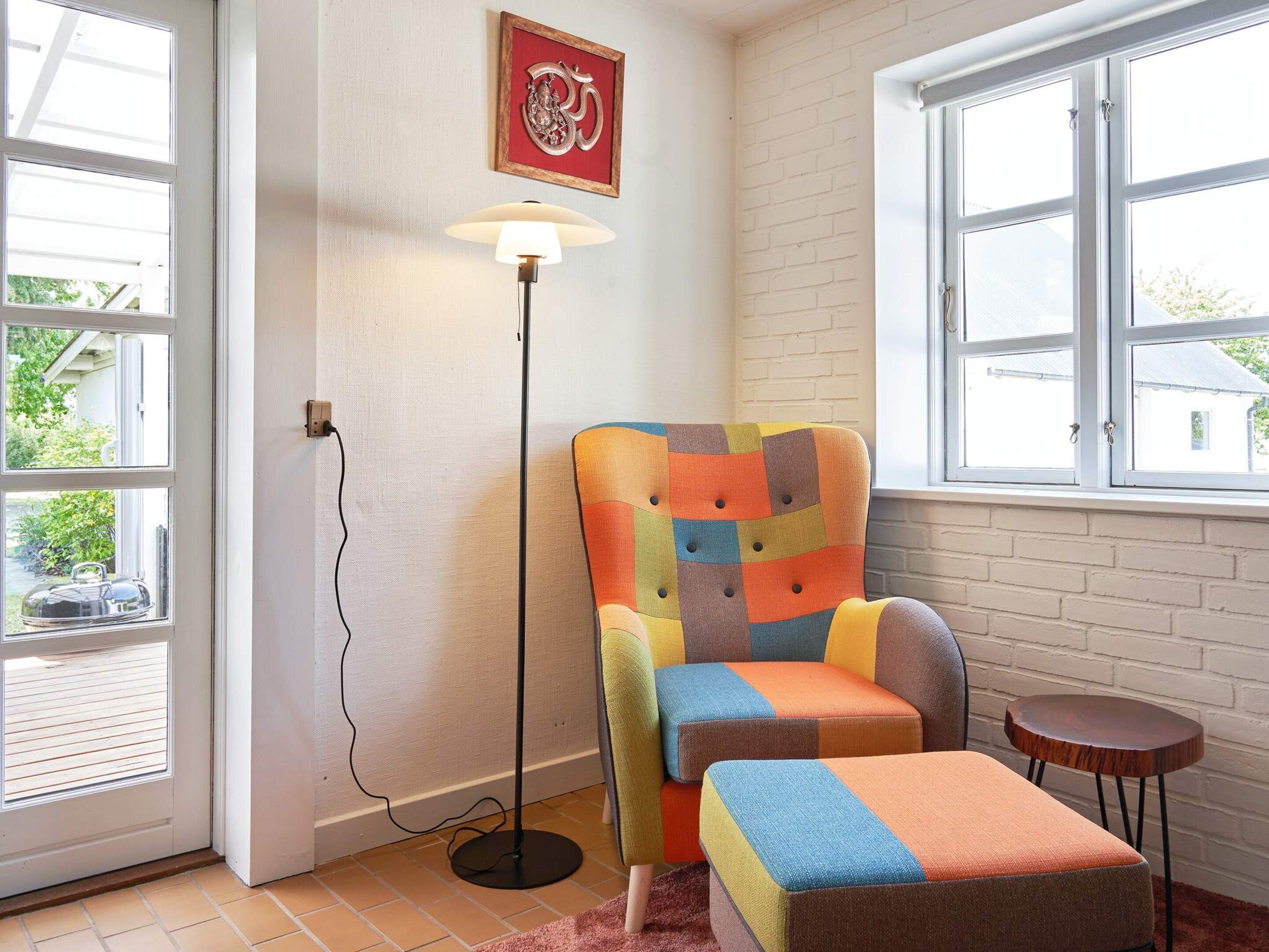 A room with a colorful armchair, a floor lamp, a small table, and a window with a view of greenery outside.