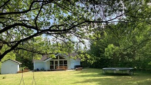 Un jardin avec une balançoire, un trampoline, un abri de jardin et une maison dotée d’une grande fenêtre.