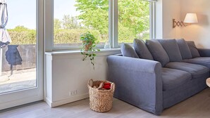 Una sala de estar moderna con un sofá gris, ventanas amplias con vistas a la vegetación y una cesta tejida en el suelo.