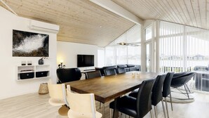 A modern dining area with a long wooden table, black and beige chairs, a television, and a large window with blinds. - 6 Person Holiday Home in Vaeggerlose (Vaeggerlose)