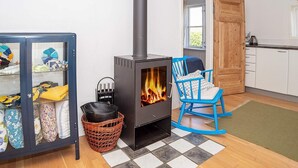 A cozy room with a wood-burning stove, a blue rocking chair, and a glass display cabinet with textiles. - 6 Person Holiday Home in Lemvig (Lemvig)