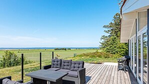 An outdoor deck with a sofa set and a table, overlooking a grassy field and the sea.