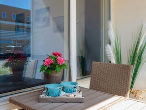 A balcony with a wicker chair, a potted plant with pink flowers, and two ceramic cups on a tray.