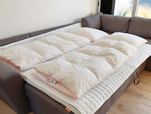 A bed with a white mattress and pillows, a grey sofa, and a wooden floor.