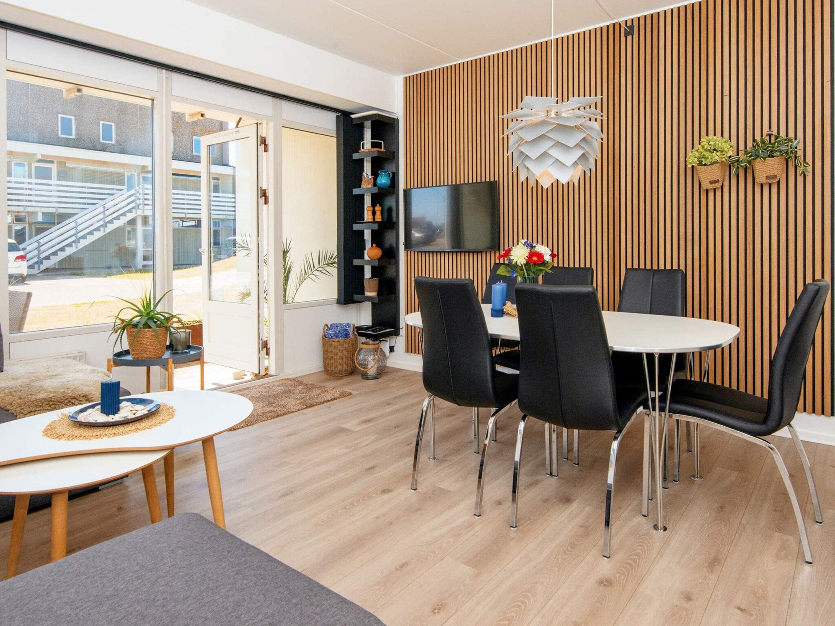 A modern dining area with a round table, black chairs, a TV mounted on a wooden wall, and a view of the outside.