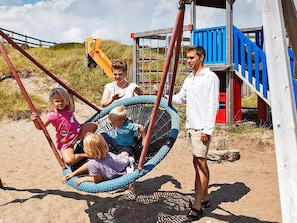 A beach scene with a swing set, a wooden structure with blue and red accents, and a sandy ground.