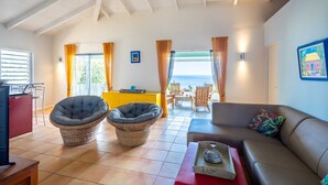 A living room with a sofa, two armchairs, a dining table with chairs, a TV, and a kitchen area with a refrigerator. - Coral Vista: Ocean View & Pool Paradise (Rif St Marie)