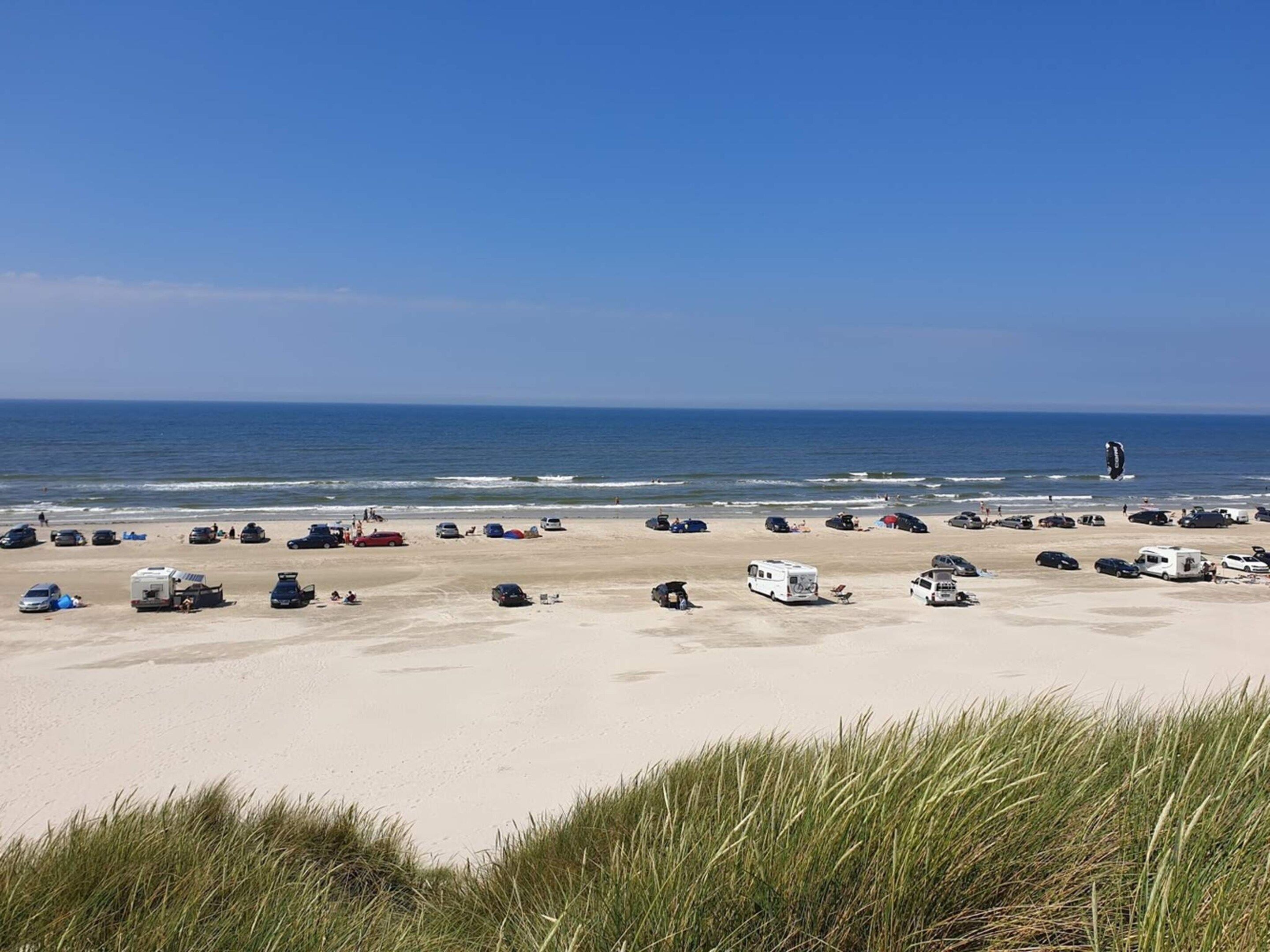 Ein Strand mit mehreren Fahrzeugen, die im Sand geparkt sind, einem klaren Himmel und dem Ozean im Hintergrund.