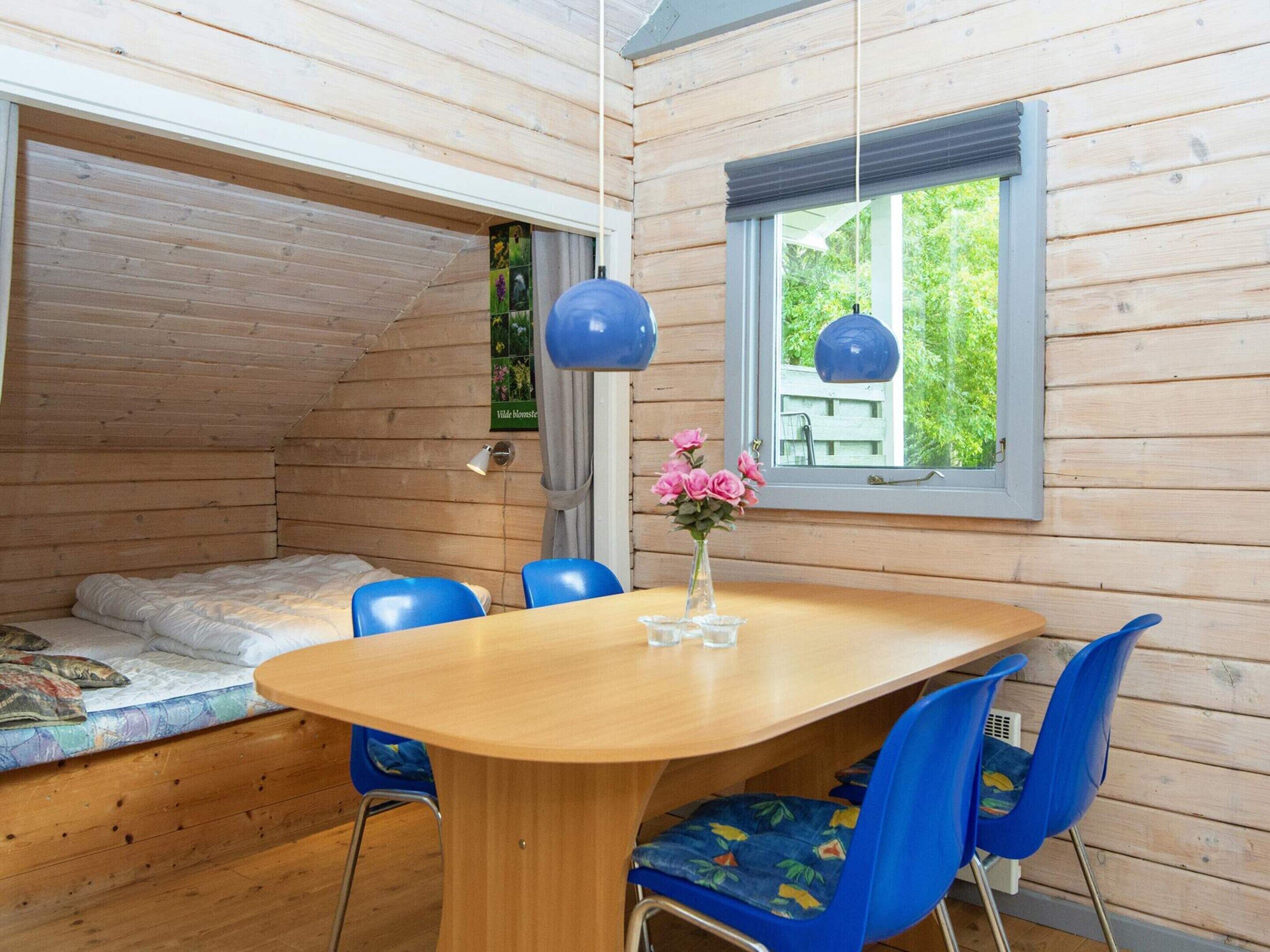 A cozy cabin interior with a wooden dining table, blue chairs, a small window with a view of greenery, and a bed with a colorful quilt.