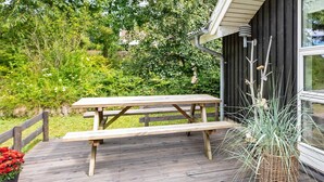 Une terrasse en bois avec une table de pique-nique, entourée de verdure et bordée d’un bâtiment doté de grandes fenêtres.