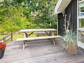 A wooden deck with a picnic table, surrounded by greenery and a building with large windows. - 6 Person Holiday Home in Fjerritslev (Fjerritslev)