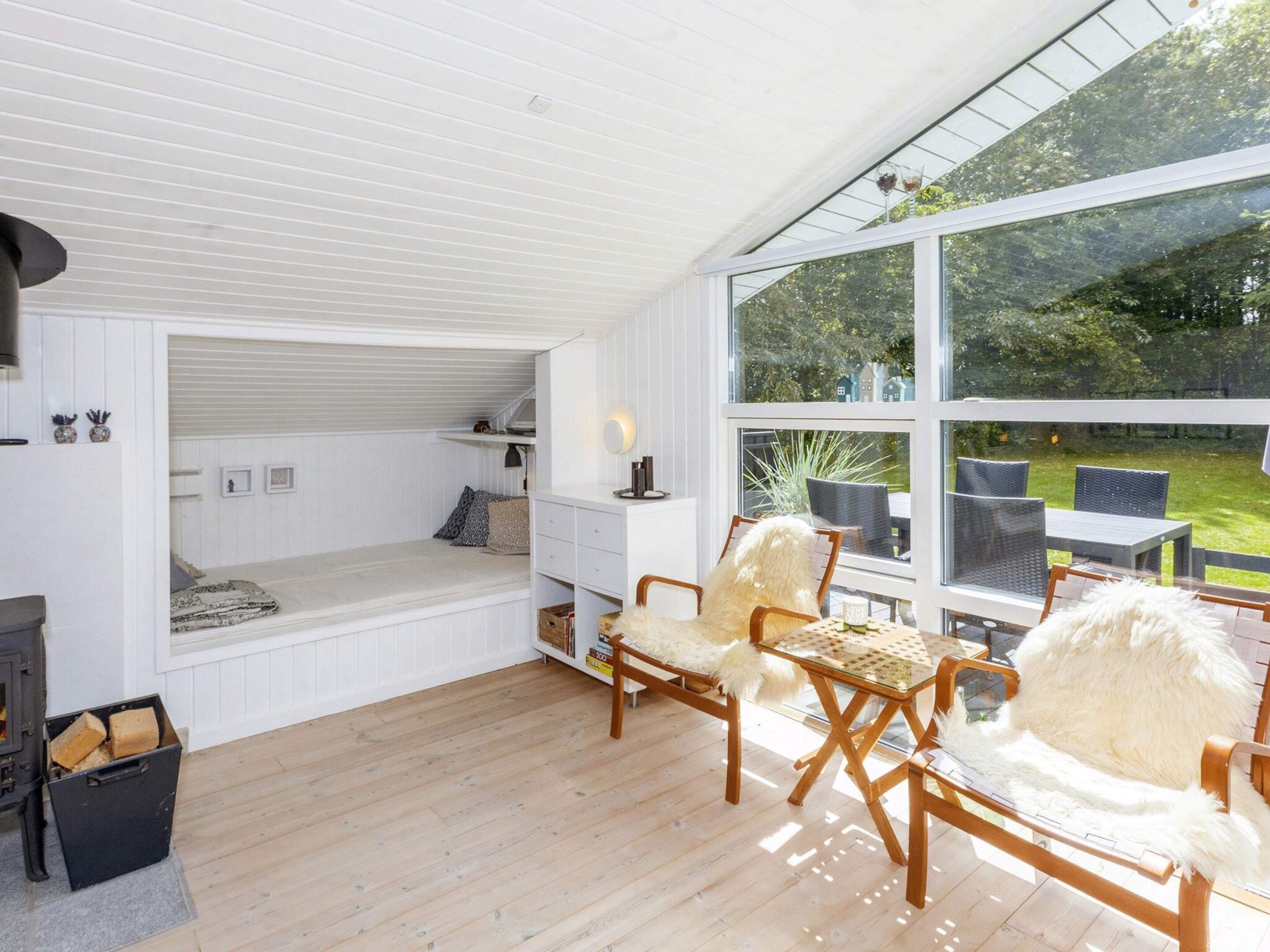 Ein Zimmer mit einem eingebauten Bett, einem Kamin, einer Essecke mit Stühlen und großen Fenstern mit Blick ins Freie.