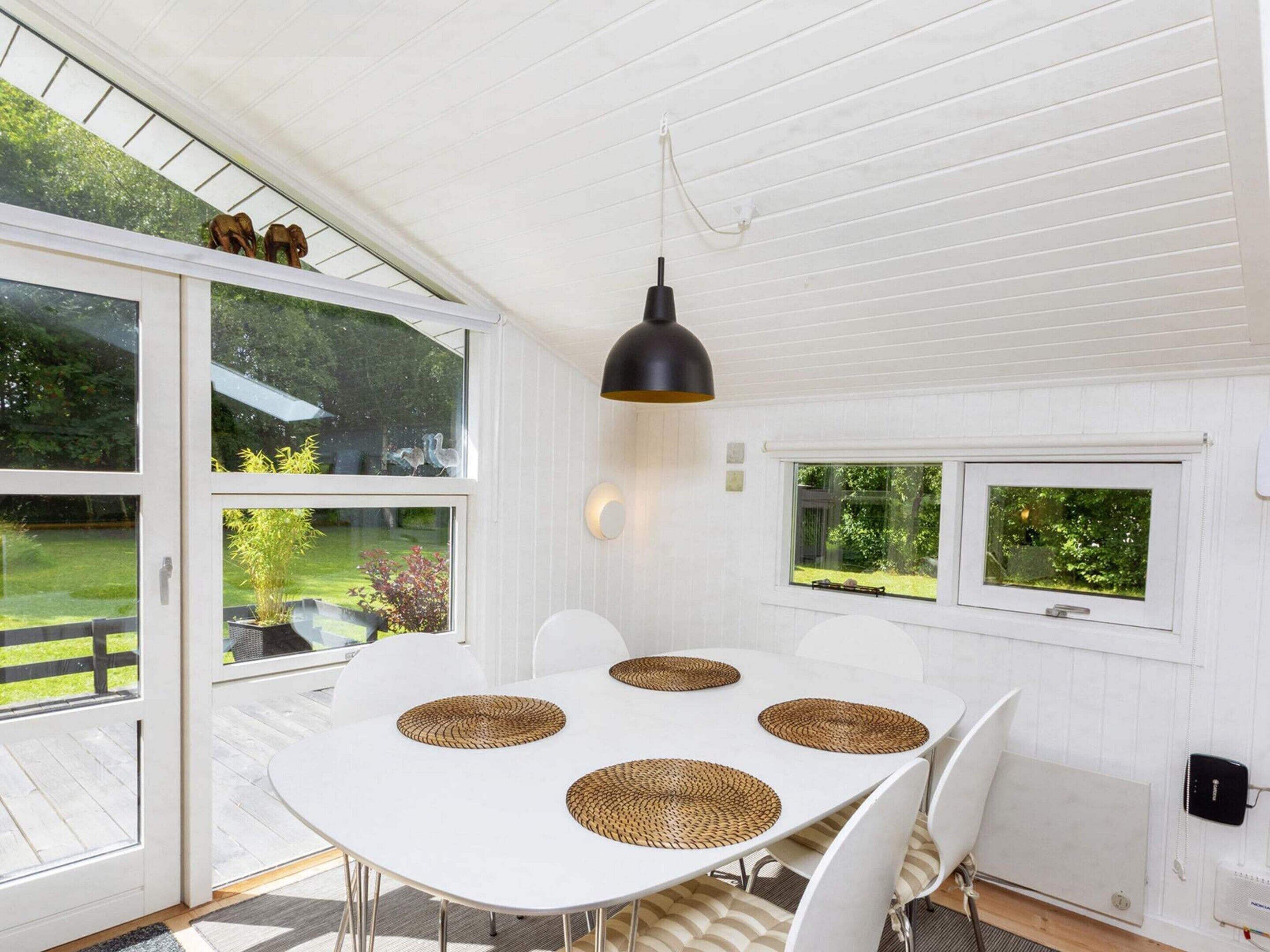 A white dining table with four woven placemats, white chairs, a black pendant light, and a view of a green garden through large windows.