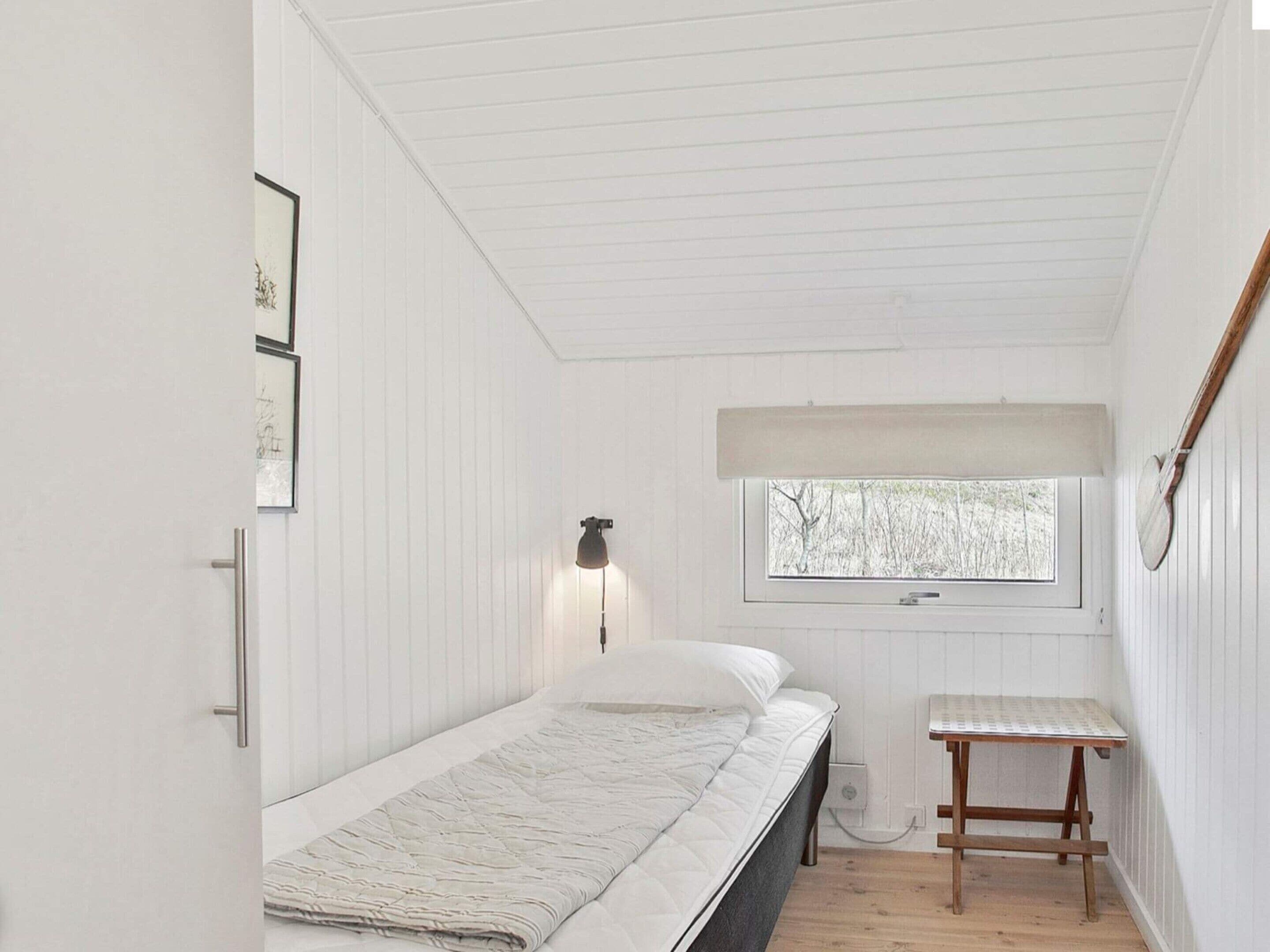 A small, white-paneled room with a single bed, a bedside table, a wall-mounted lamp, and a window with a view of trees.
