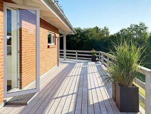 Une terrasse en bois avec une porte vitrée donnant sur une pièce, le tout entouré d’une clôture blanche et de verdure.