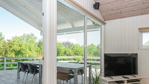 A modern living room with a large TV, a dining table with chairs, and a balcony with a view of trees. - 10 Person Holiday Home in Juelsminde-by Traum (Horsens)