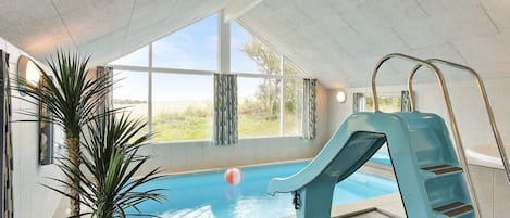 An indoor swimming pool with a blue slide, a potted plant, and large windows.
