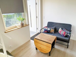 A room with a sofa, a wooden table, and a window with a view of outdoor furniture and greenery.