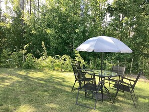 An outdoor dining area with a round table and chairs, surrounded by a grassy lawn and dense foliage.