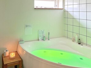 A bathroom with a green-lit bathtub, a small wooden side table with a candle, and a window above the tub. - 3 Person Holiday Home in Ebeltoft (Ebeltoft)